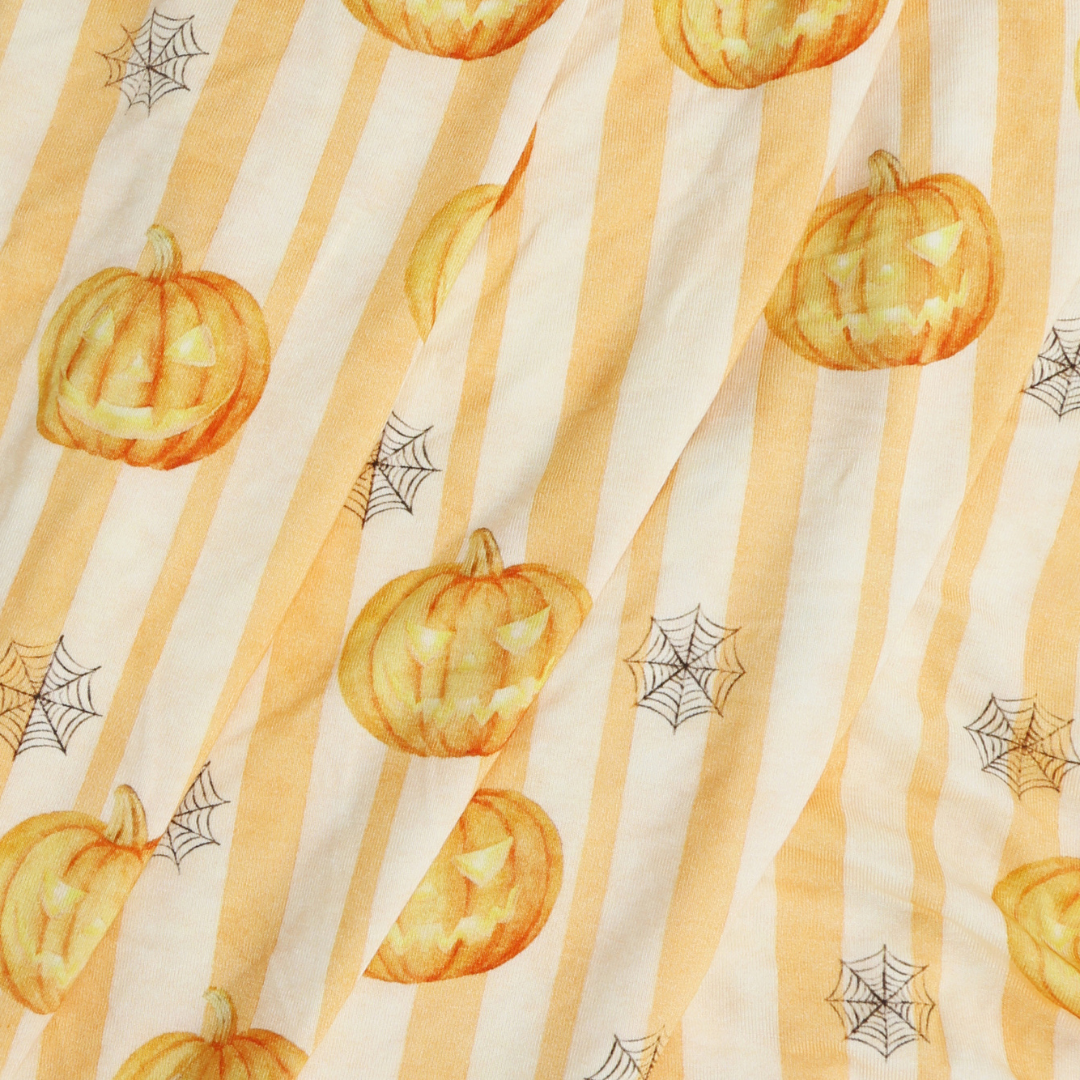 A close-up of a fabric surface with vertical cream and pale yellow stripes, decorated with orange jack-o'-lantern pumpkins and black spiderweb motifs in a Halloween pattern.
