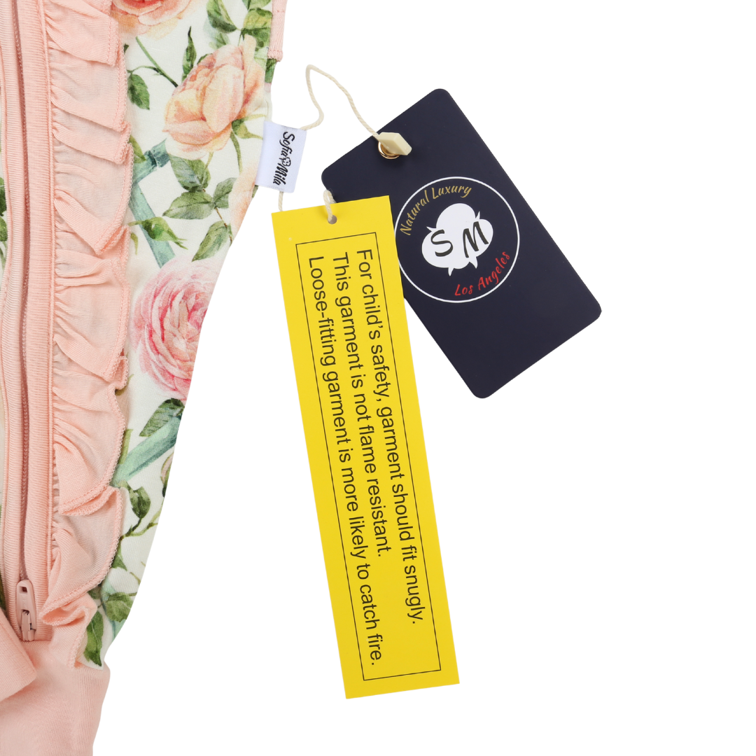 A close-up of a garment tag and care label attached to a pink floral ruffled garment. The tag is dark navy blue with a circular white logo reading 'Safeway of Loving SM Care Label', and the yellow safety tag reads 'For Child's Safety Garment should it snugly This garment is not flatter easiest to loose fitting garment is more likely to catch the'.