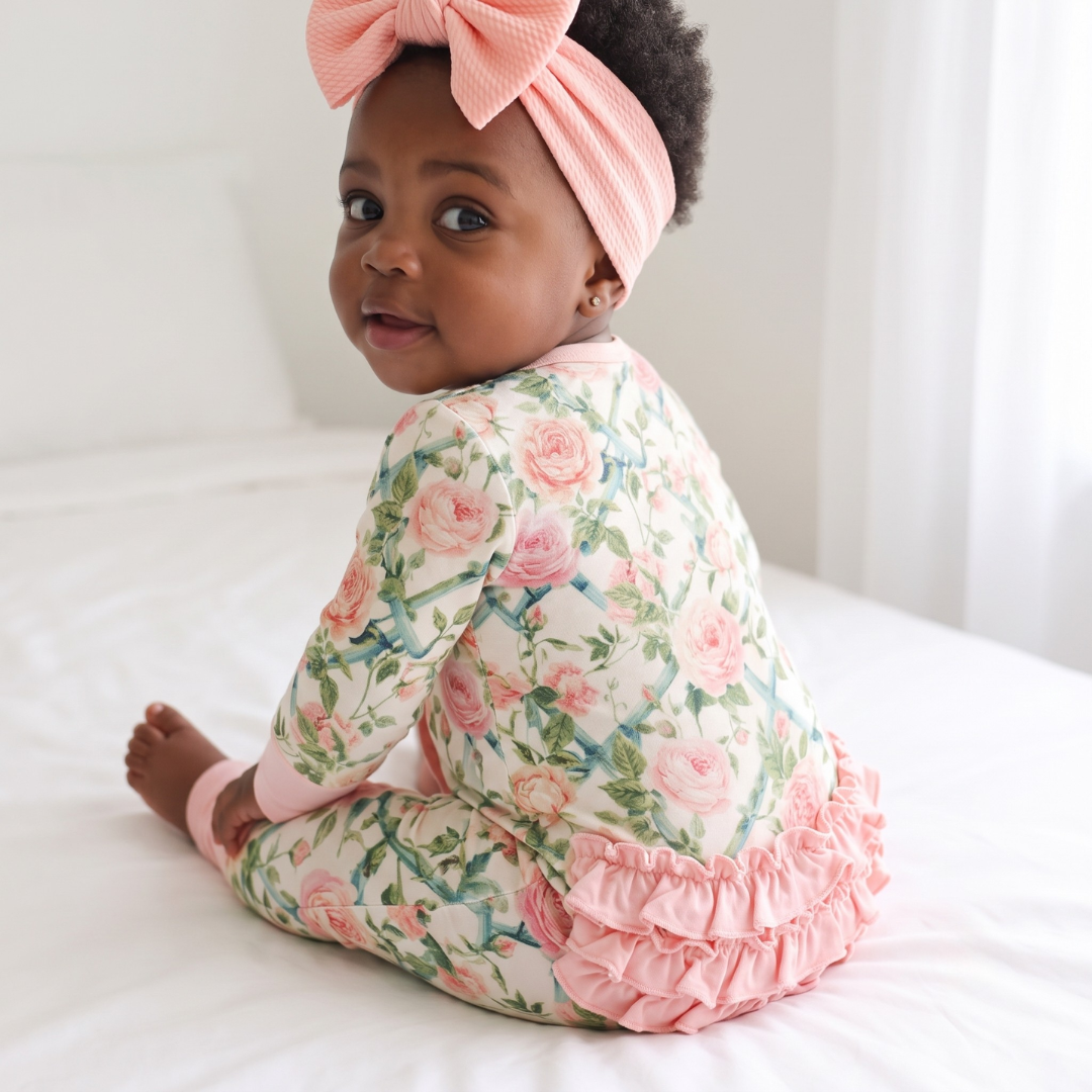 A baby girl wearing a pink and green floral print long-sleeve romper and a matching pink bow headband, sitting on a white bed.