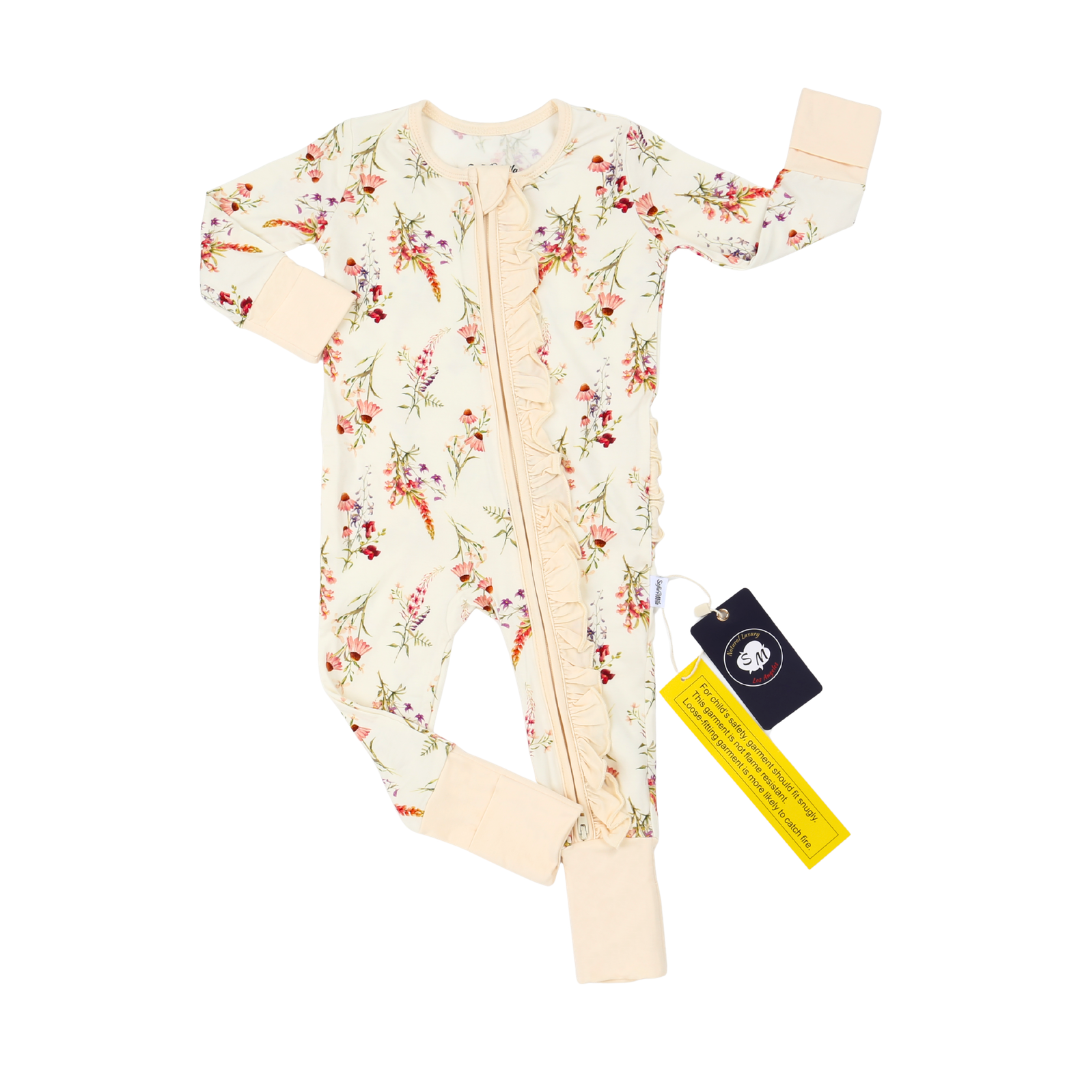 A baby long-sleeve footless sleeper in cream with a delicate multicolor floral print, featuring a ruffle down the front zipper and light peach cuffs and leg cuffs. A black and yellow brand tag is attached.