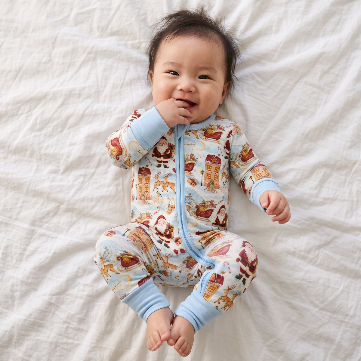 A baby lying on a white textured blanket, wearing a long-sleeve zip-up sleeper with a light blue trim and a festive Santa Claus and holiday print.
