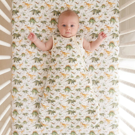 A baby lying on their back inside a crib, wearing a sleeveless swaddle blanket with a green and yellow dinosaur print on an off-white background. The matching crib sheet also features the same dinosaur pattern.