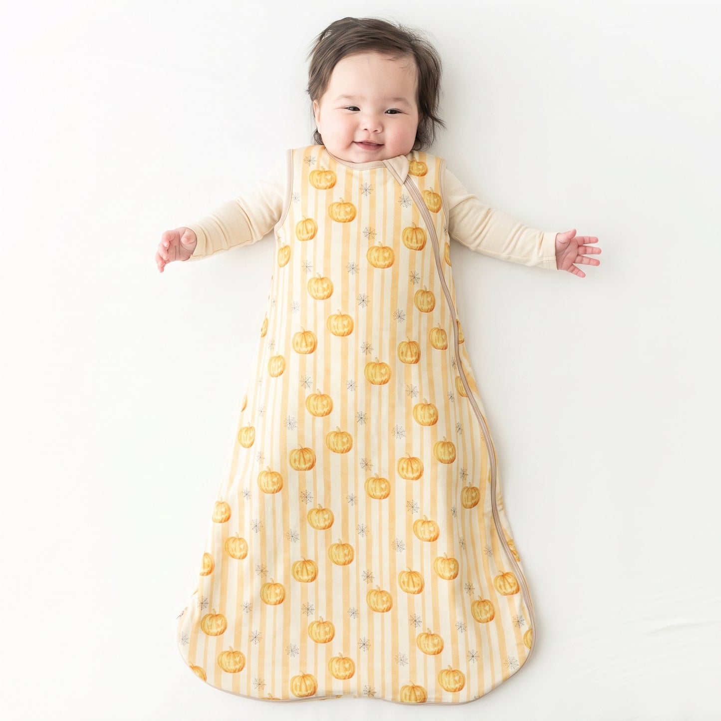 A baby lying on their back wearing a long-sleeve sleep sack with a yellow and white striped pattern and orange pumpkin print, on a white background.