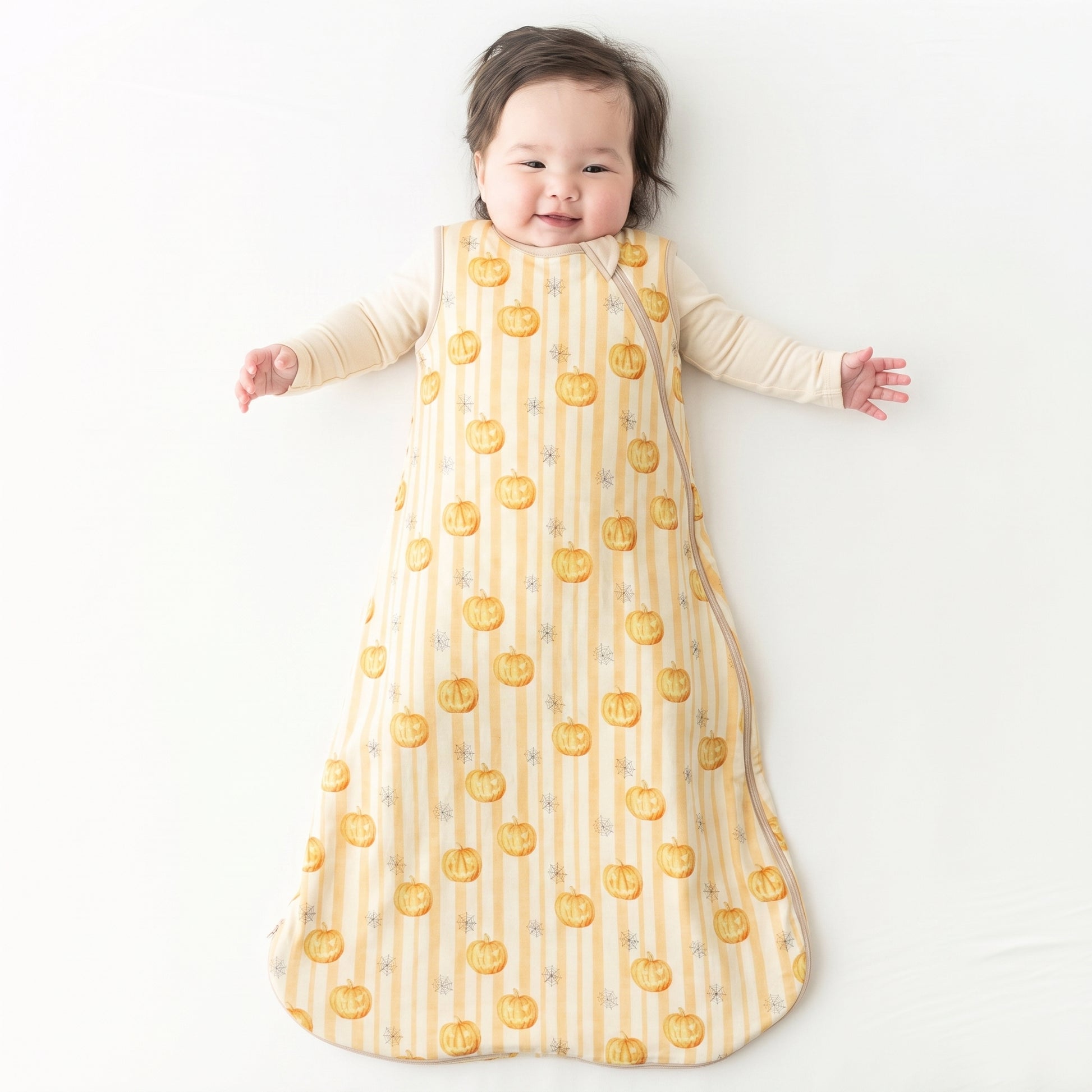 A baby lying on their back wearing a long-sleeve sleep sack with a yellow and white striped pattern and orange pumpkin print, on a white background.