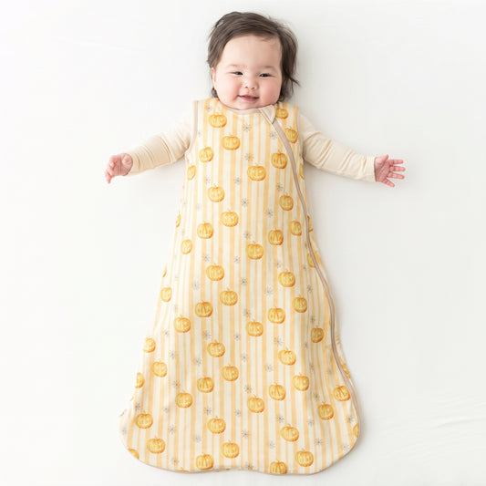 A baby lying on their back wearing a long-sleeve sleep sack with a yellow and white striped pattern and orange pumpkin print, on a white background.