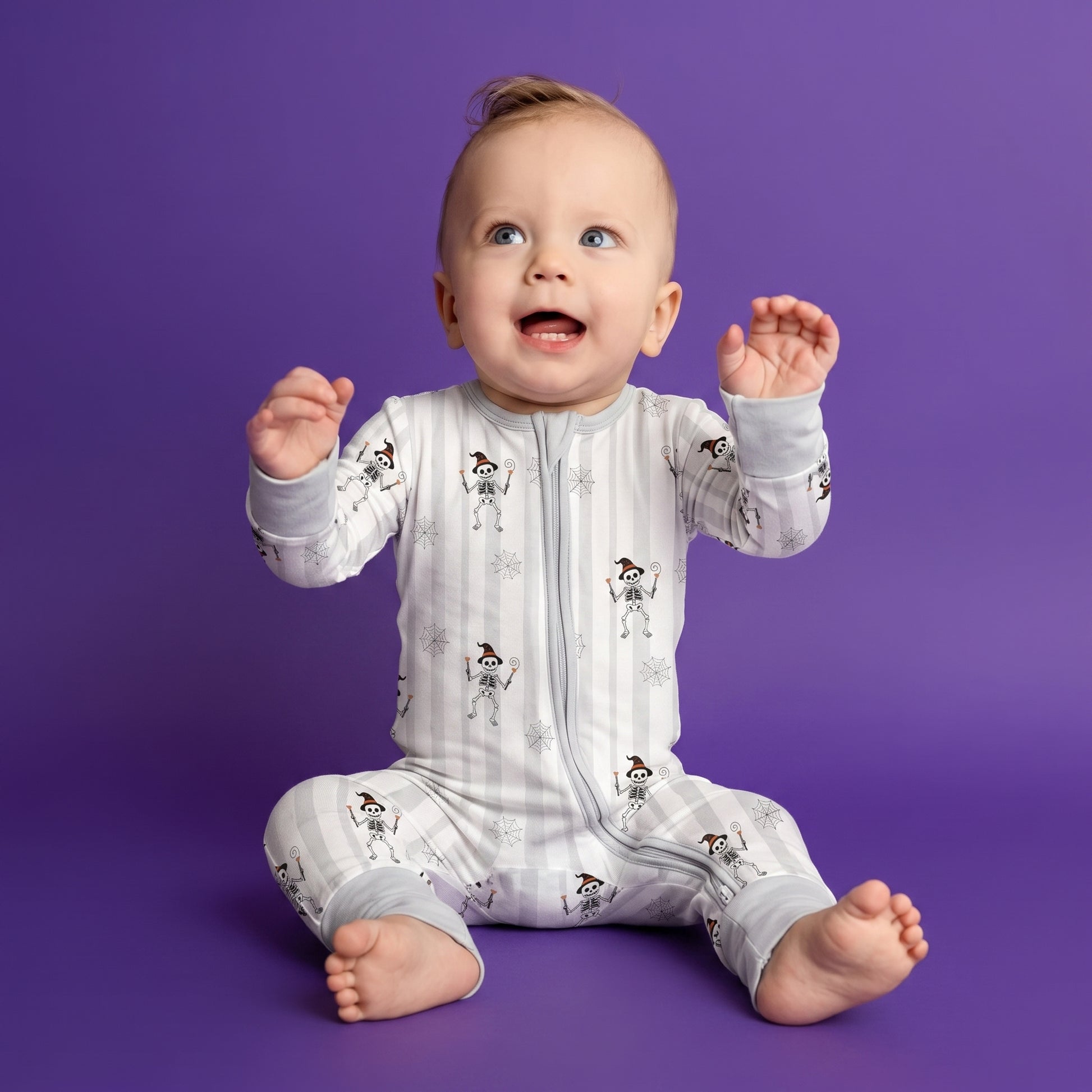 A baby sitting on a purple studio background wearing a light gray and white zip-up sleeper with a snowman pattern.