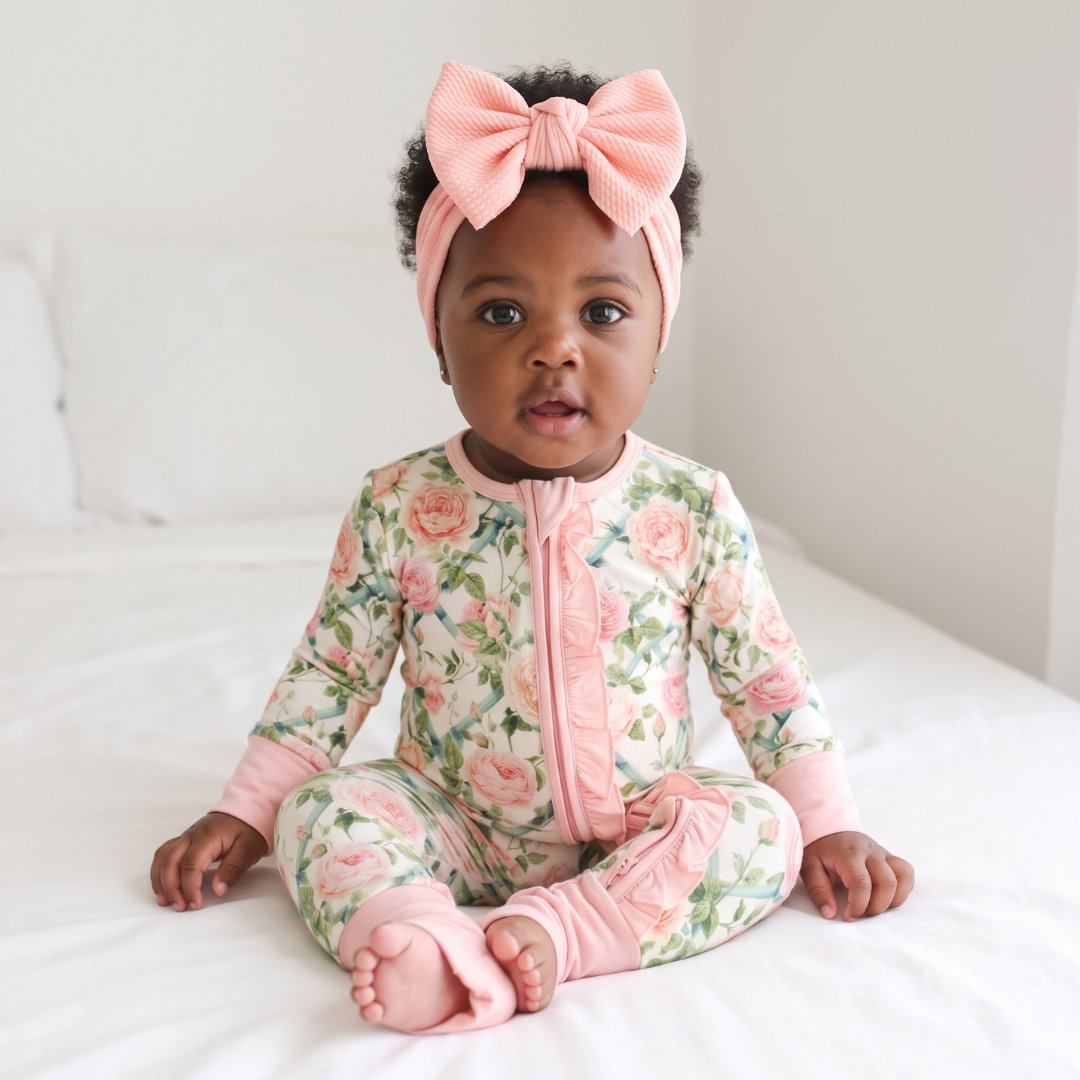 A baby sitting on a white bed wearing a long-sleeve floral zip-up onesie with pink trim and a matching pink bow headband.