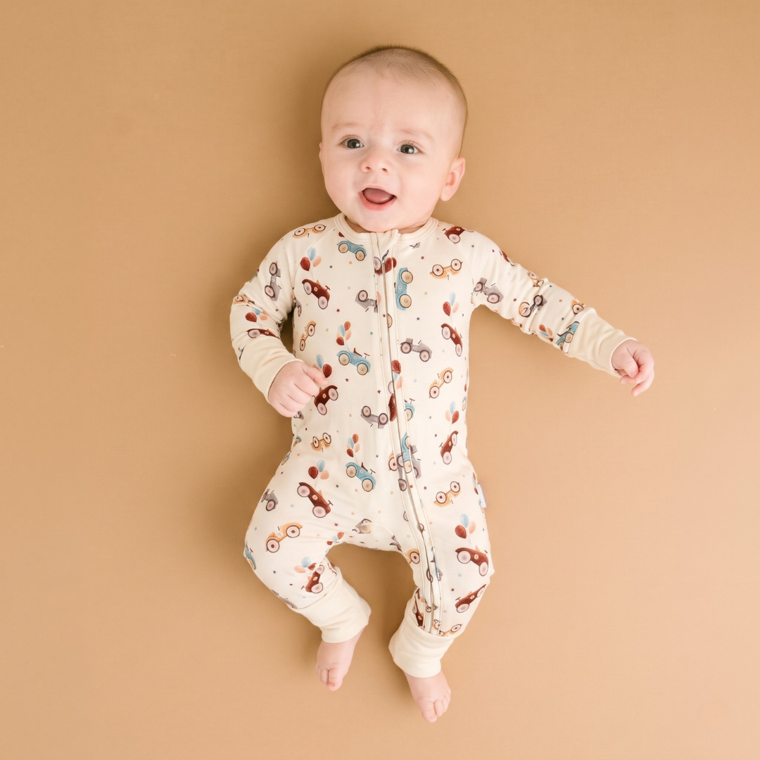 A baby wearing a cream-colored footless sleeper with a colorful bicycle pattern, lying on a solid tan background.