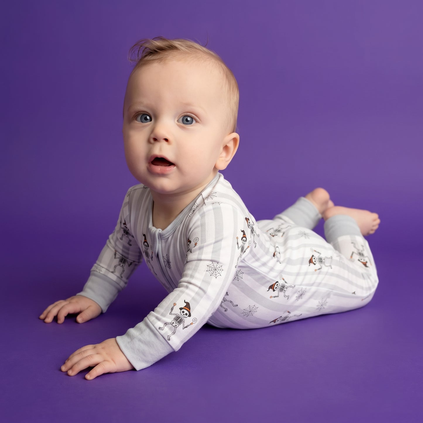 A baby wearing a light gray and white striped long-sleeve footless pajama romper with a small dog print, crawling on a solid purple studio background.