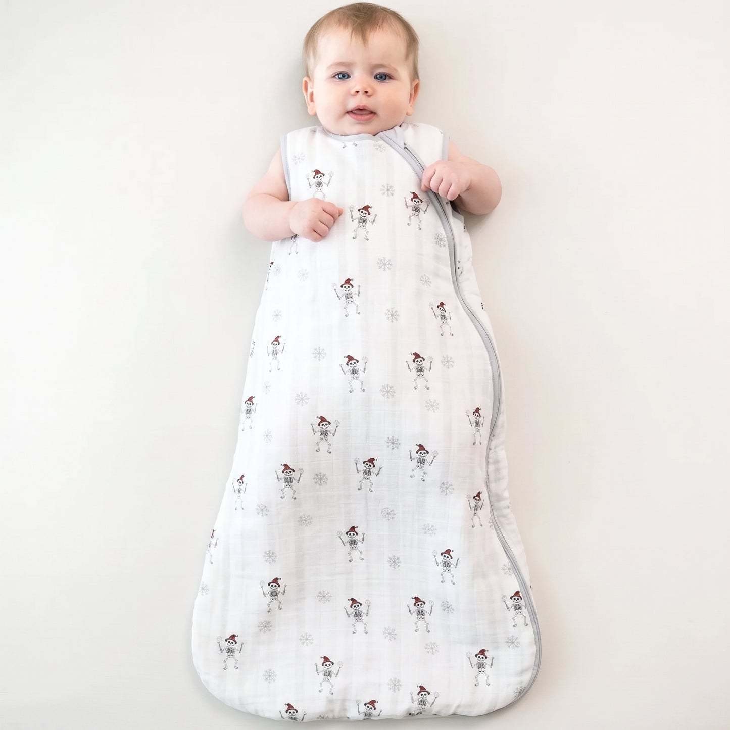 A baby wearing a white muslin sleep sack with a snowman pattern, lying on a light cream background.