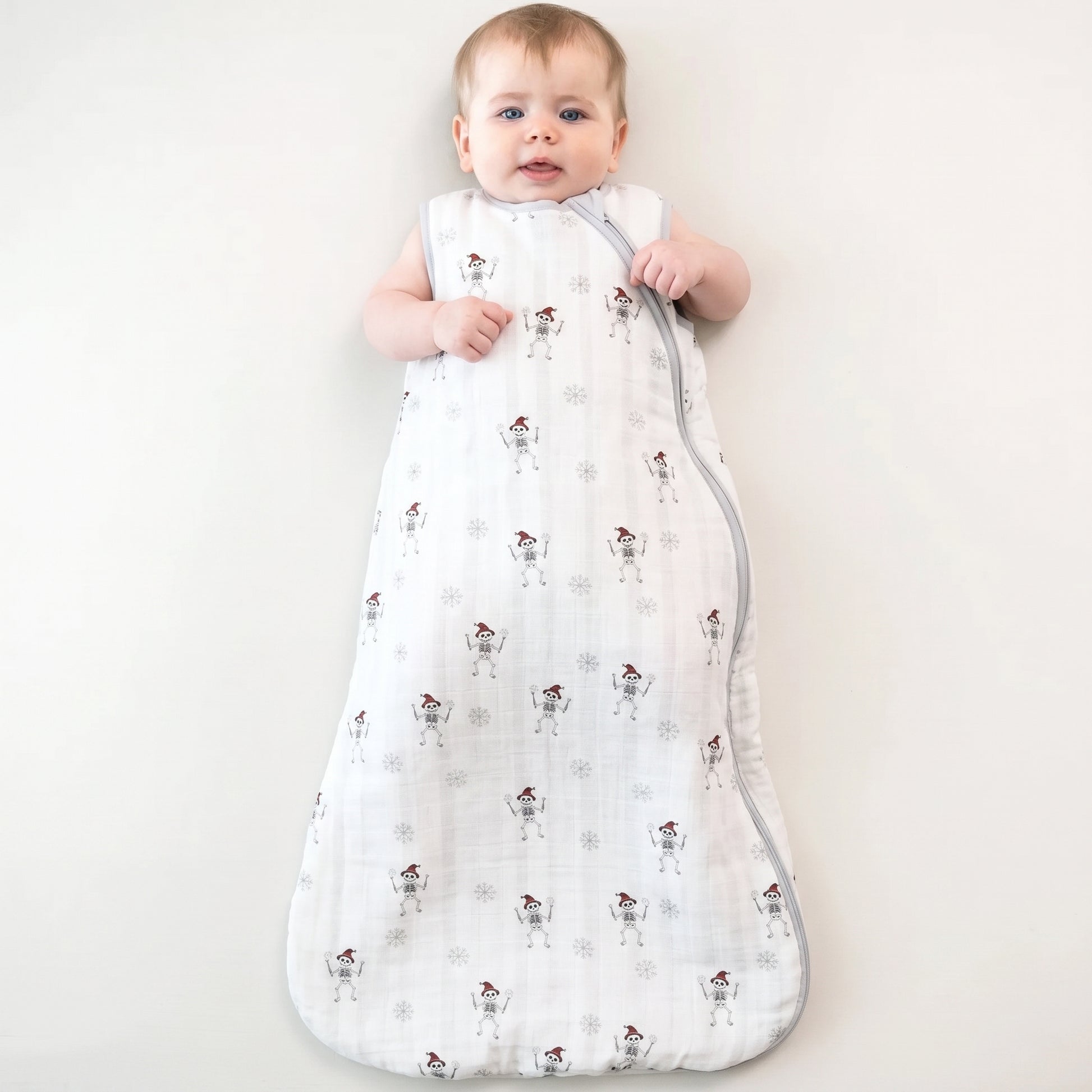 A baby wearing a white muslin sleep sack with a snowman pattern, lying on a light cream background.