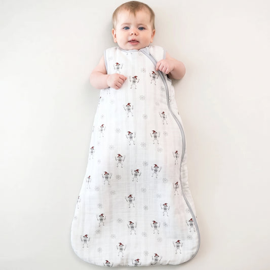 A baby wearing a white muslin sleep sack with a snowman pattern, lying on a light cream background.