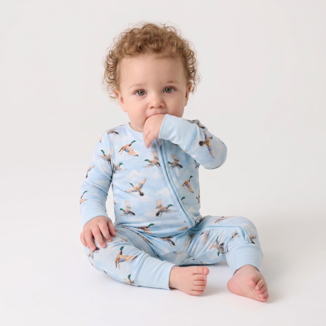 A baby with curly light brown hair sits on a white studio floor wearing a light blue long-sleeve footless sleeper with a zipper and an all-over print of flying ducks.