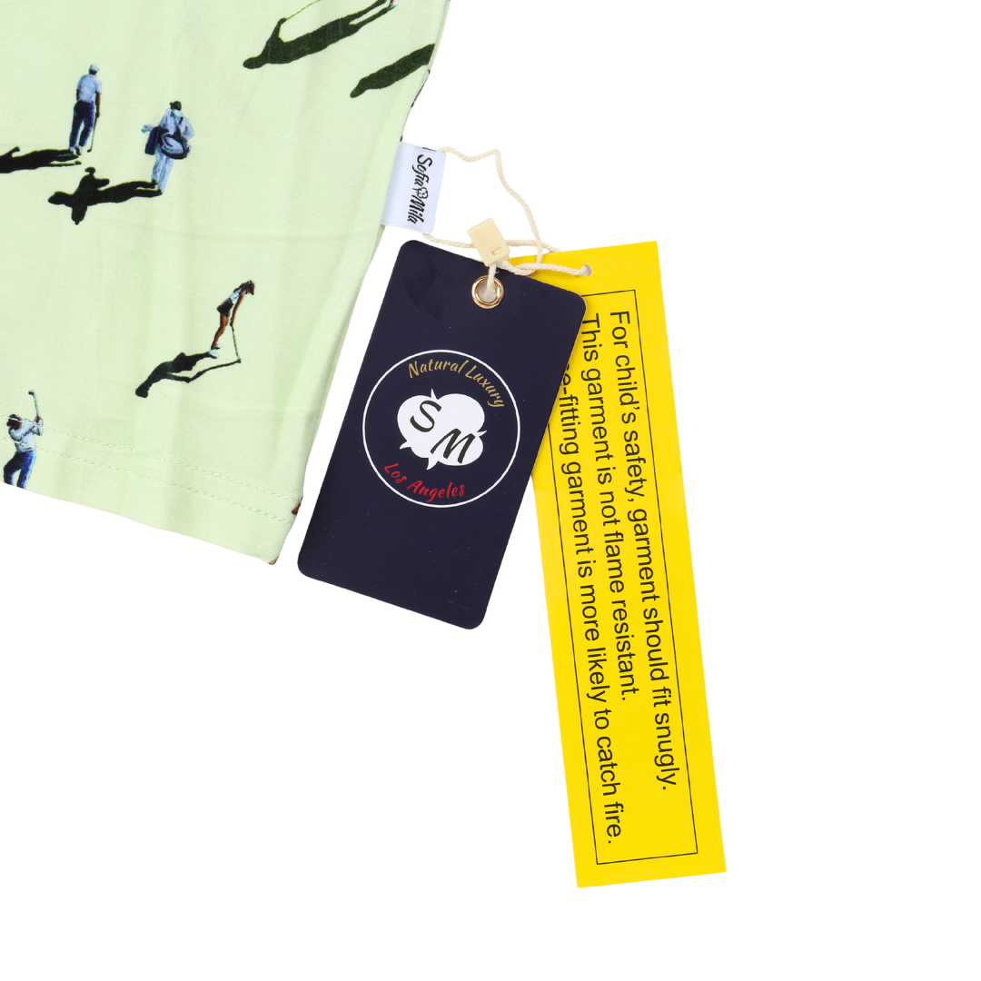 A close-up of a light green children's garment with a black golf-themed print, featuring a dark navy safety patch and a bright yellow care/information tag on a white background.