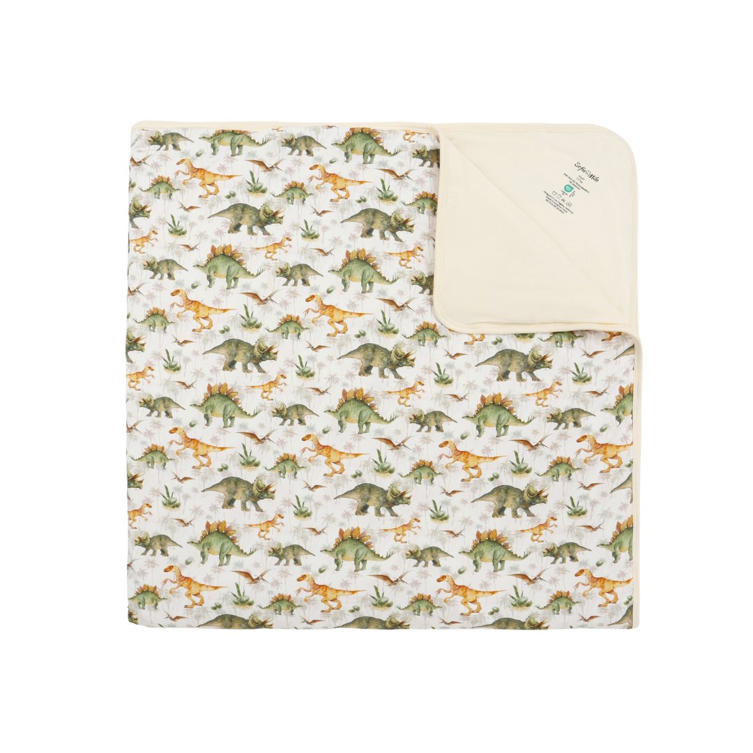 A folded baby blanket featuring a colorful dinosaur pattern on an off-white background, with a solid cream-colored reverse side showing a small printed label.
