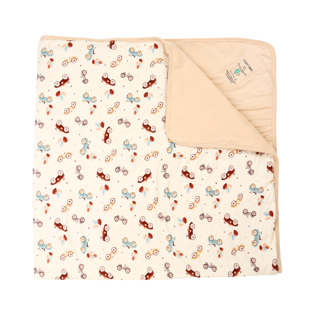 A folded baby blanket with a cream background and a playful pattern of small bicycles in brown, light blue, and rust red. The underside is solid cream with a soft texture and a small sewn-in label.