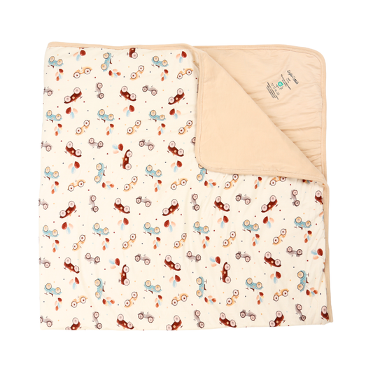 A folded baby blanket with a cream background and a playful pattern of small bicycles in brown, light blue, and rust red. The underside is solid cream with a soft texture and a small sewn-in label.