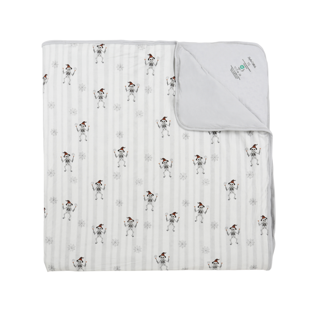 A folded muslin blanket with a white and gray striped background, featuring a repeating pattern of cartoon skeletons wearing Santa hats. The top right corner is folded over to show the solid white reverse side and a small printed brand label.