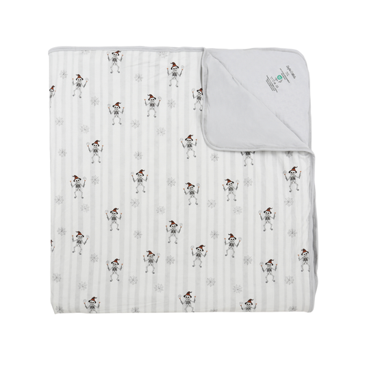 A folded muslin blanket with a white and gray striped background, featuring a repeating pattern of cartoon skeletons wearing Santa hats. The top right corner is folded over to show the solid white reverse side and a small printed brand label.