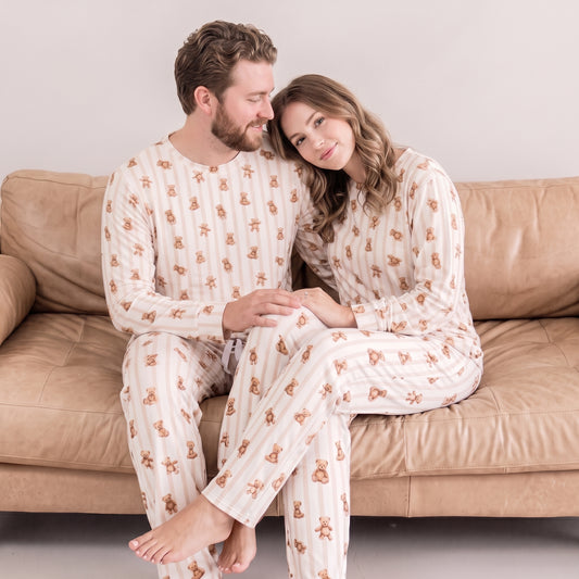 A man and a woman sitting closely together on a tan leather couch, wearing matching long-sleeve pajamas with vertical cream stripes and a repeating brown teddy bear print.