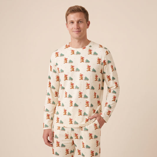 A man wearing a long-sleeve pajama shirt and matching pants with an all-over Christmas print of brown bears and green pine trees on a cream background, set against a solid beige studio backdrop.