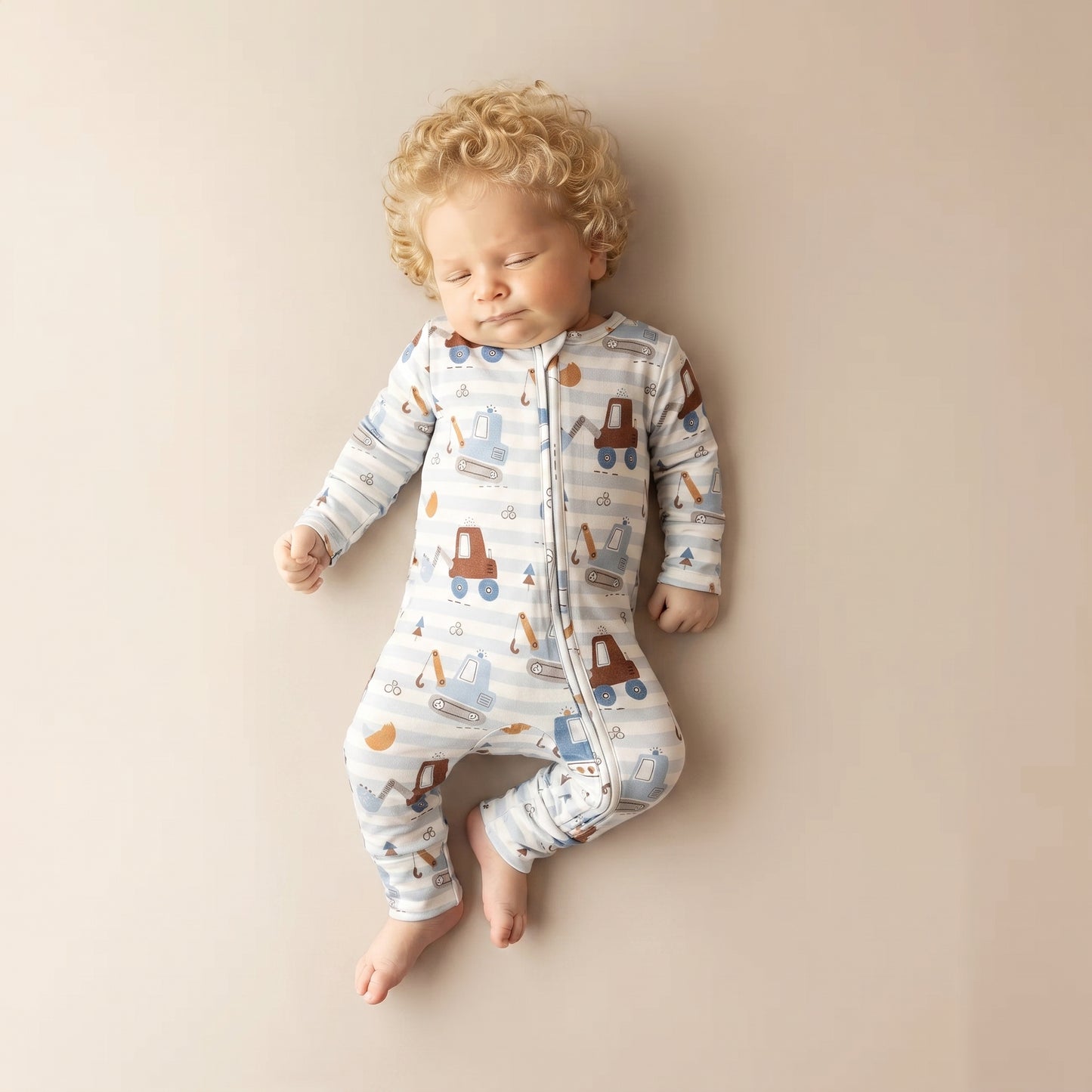 A sleeping baby with curly blond hair wearing a long-sleeve zip-front sleeper with a blue, white, and brown construction truck pattern, lying on a beige background.