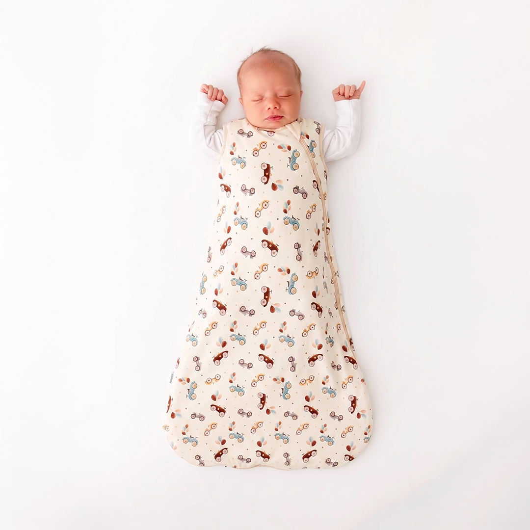 A sleeping newborn baby wearing a white long-sleeve shirt and swaddled in a cream-colored sleep sack with a colorful pattern of small cartoon figures, lying on a solid white background.