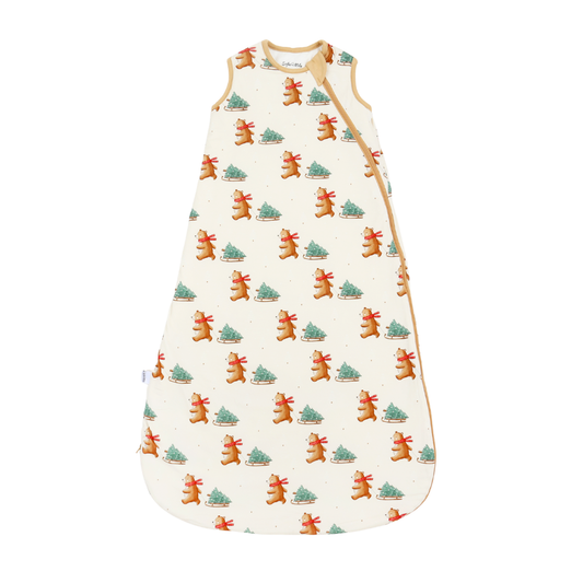 A sleeveless baby sleep sack with a festive Christmas print featuring gingerbread men and Christmas trees on an off-white background, with tan trim and side snaps.
