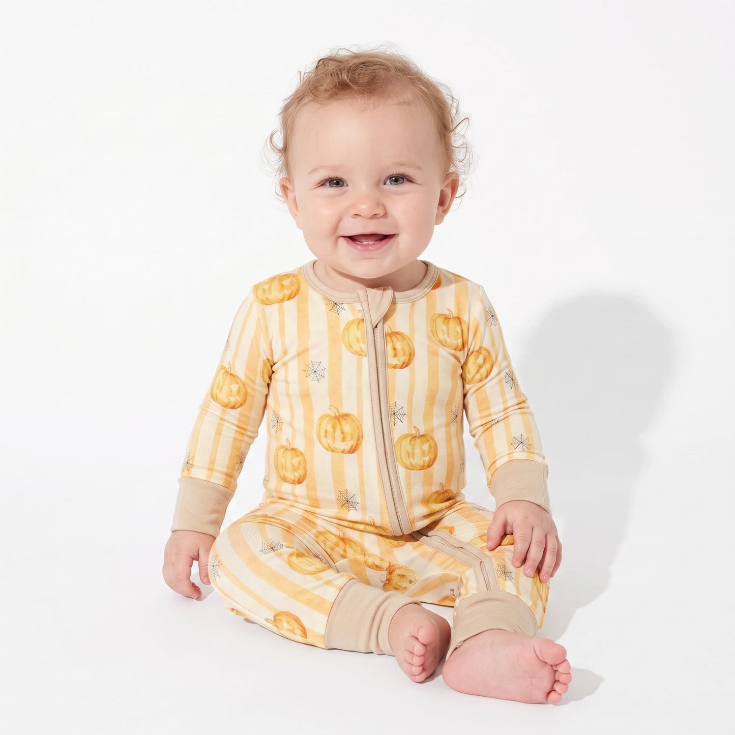 A smiling baby sitting on a white studio background, wearing a long-sleeve footless zip-up pajama in pale yellow and beige stripes with orange jack-o'-lantern prints.