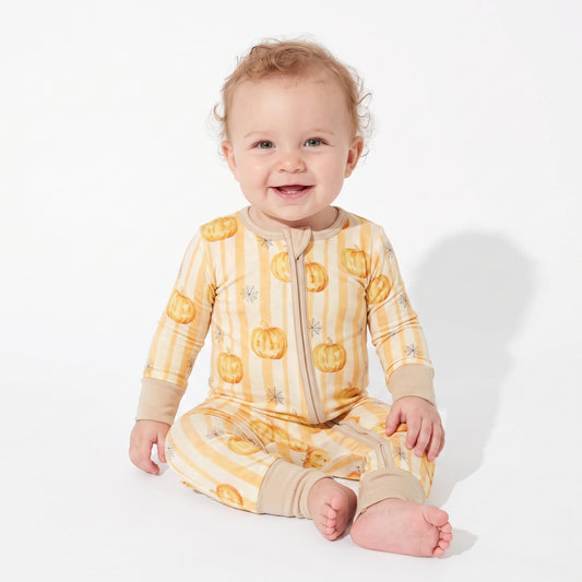 A smiling baby sitting on a white studio background, wearing a long-sleeve footless zip-up pajama in pale yellow and beige stripes with orange jack-o'-lantern prints.