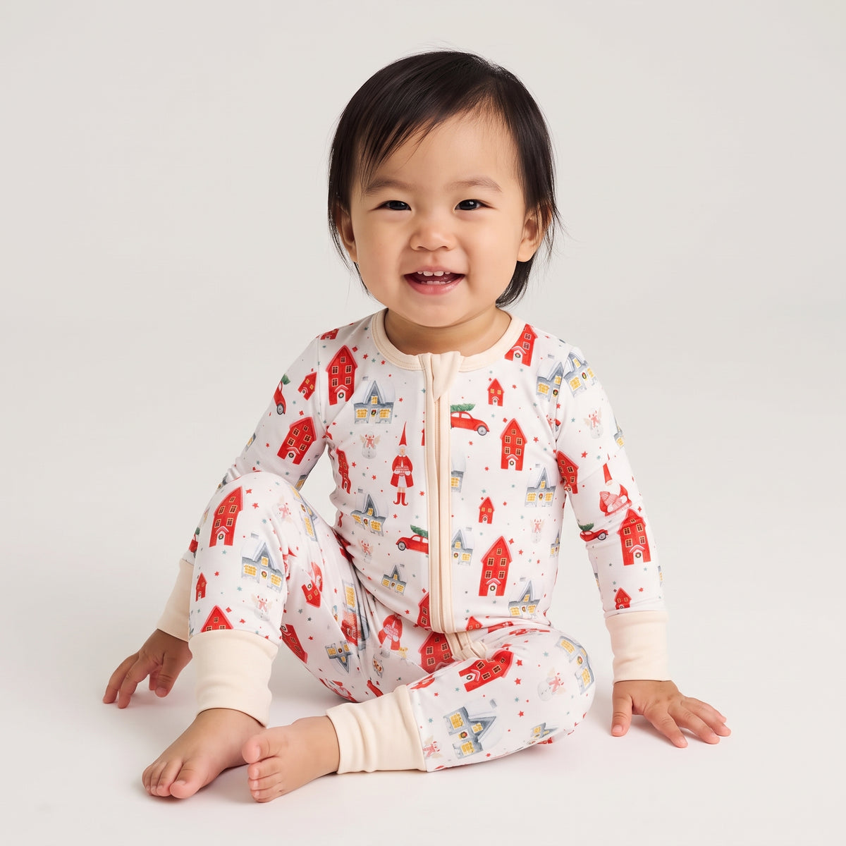 A smiling toddler sits cross-legged on a light gray studio background wearing a one-piece zip-front pajama top and matching pants with a small red, blue, and yellow house print on an off-white base.