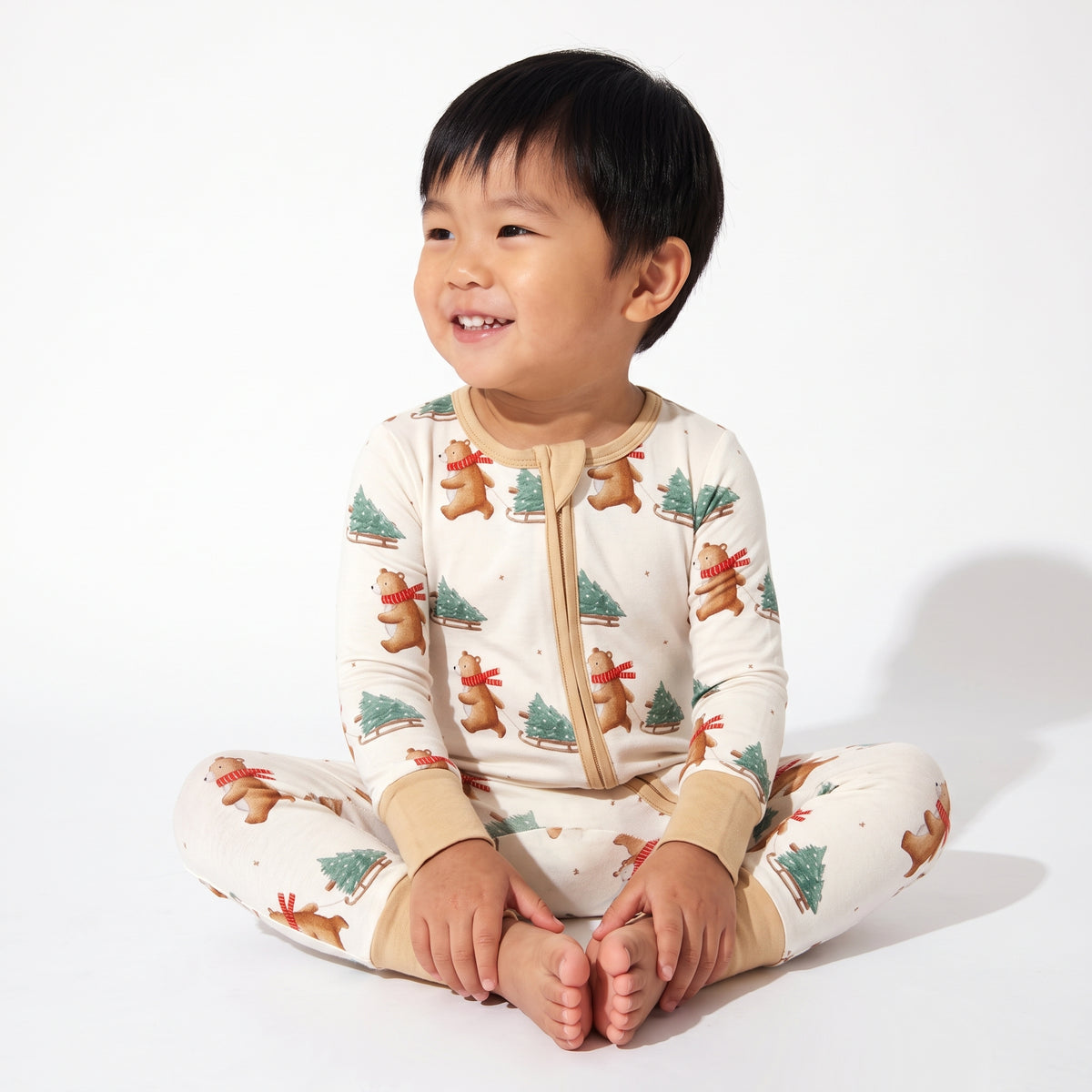 A smiling young child sitting cross-legged on a white studio background, wearing a cream one-piece pajama top and pants set with a festive print of gingerbread men, Christmas trees, and small red ornaments.