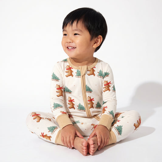 A smiling young child sitting cross-legged on a white studio background, wearing a cream one-piece pajama top and pants set with a festive print of gingerbread men, Christmas trees, and small red ornaments.