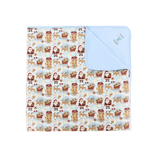 A square baby blanket with a festive Christmas print featuring Santa Claus, reindeer, sleighs, and gingerbread houses on a white background, folded to show a solid light blue reverse side with a small embroidered logo.