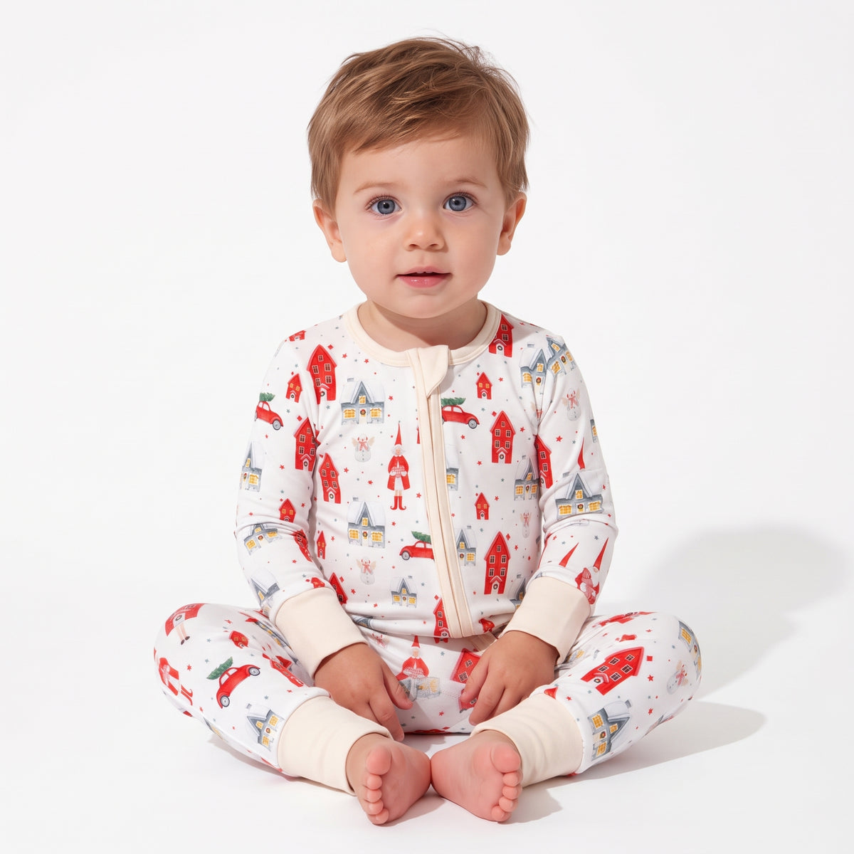 A toddler sitting cross-legged wearing a cream-colored zip-up pajama set with a red and multicolor winter village print, photographed against a clean white studio background.