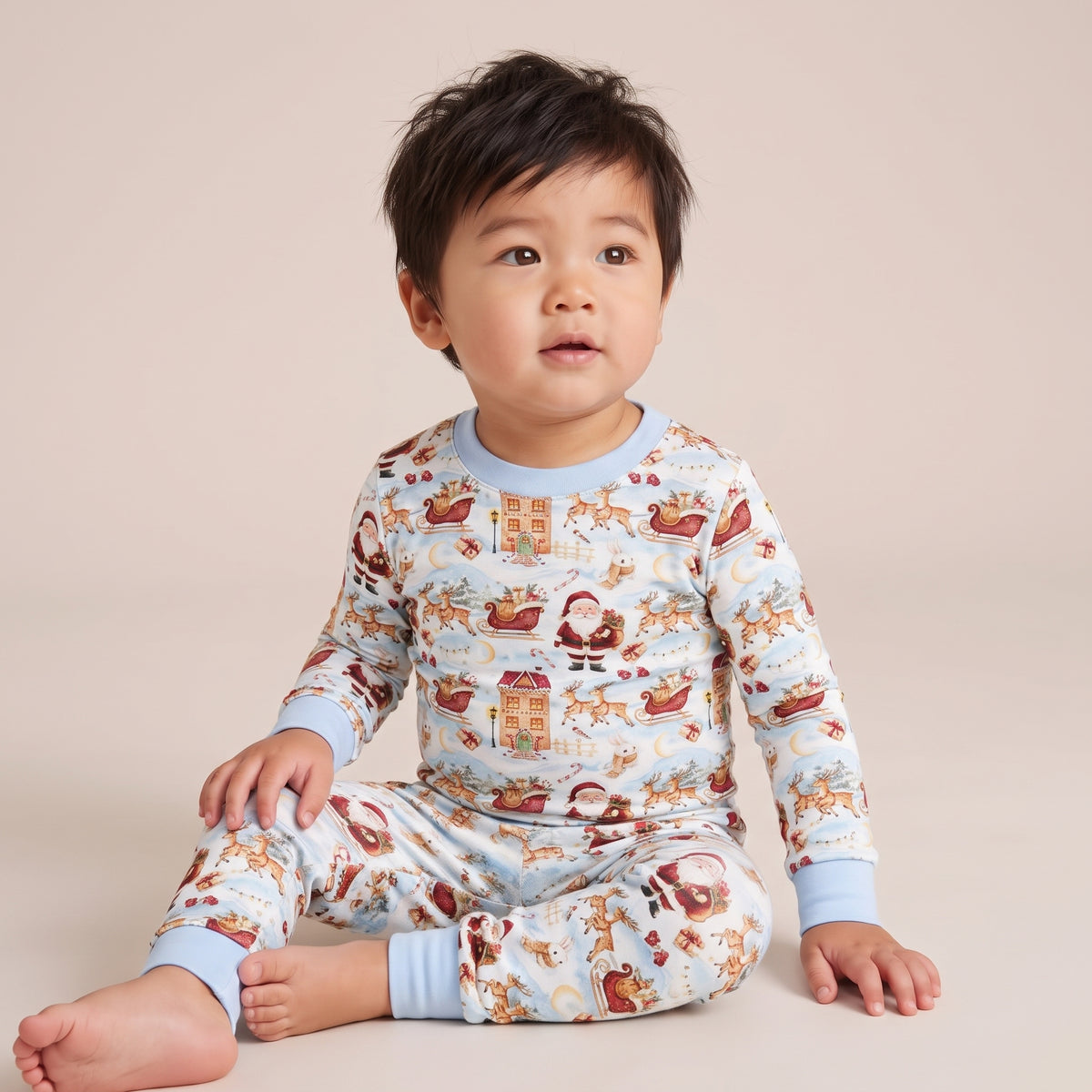 A toddler sitting cross-legged wearing a light blue long-sleeve pajama set with a festive Christmas print of Santa Claus, reindeer, sleighs, and gingerbread houses, set against a solid beige studio background.