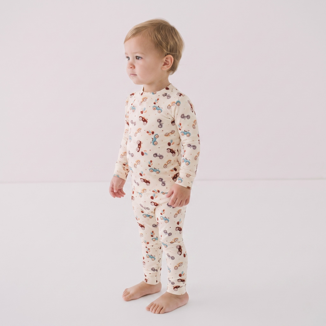 A toddler standing in profile wearing a cream-colored long-sleeve pajama set with a colorful cartoon pattern, photographed against a plain white studio background.