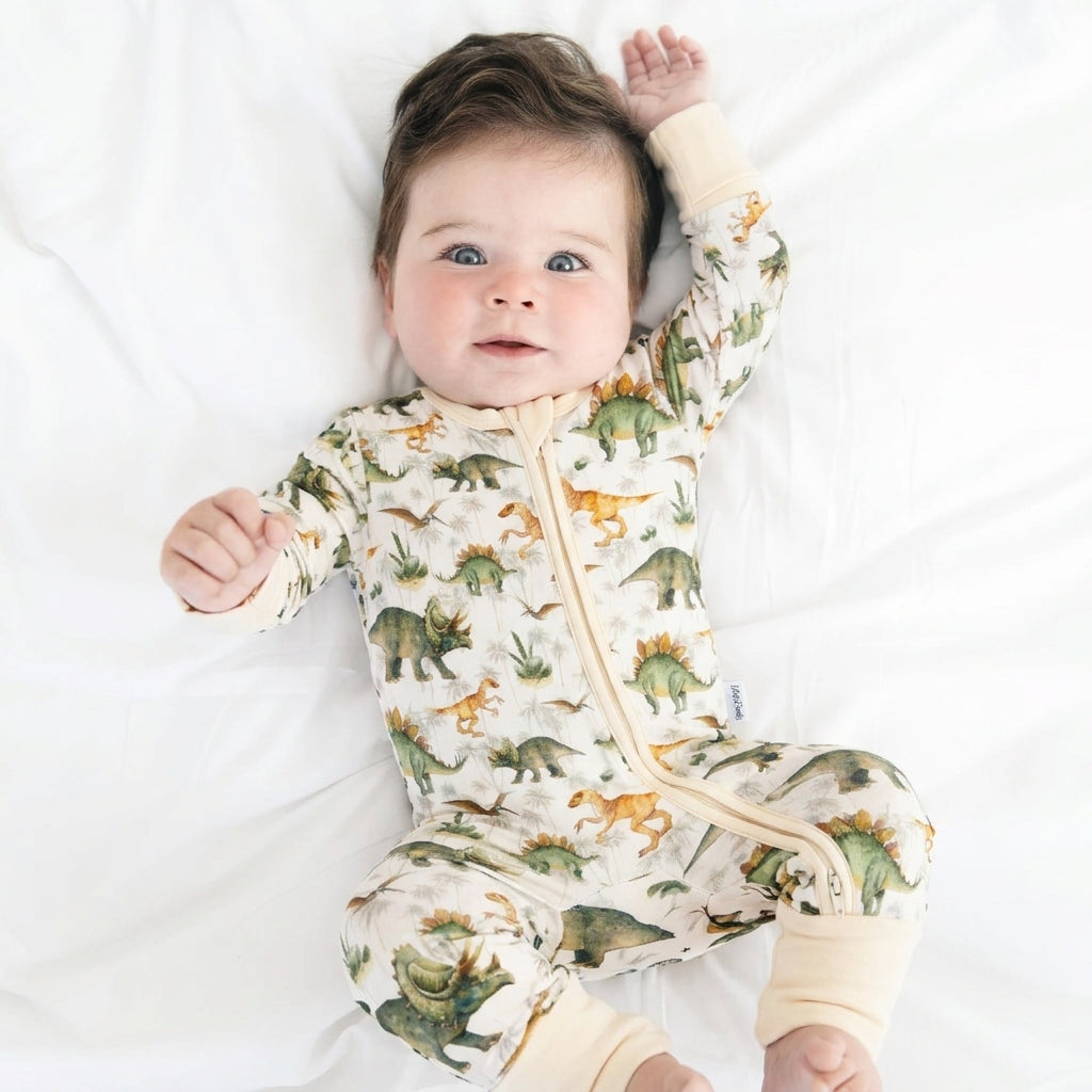 A baby lying on white bedding wearing a long-sleeve zip-front footless sleeper with an all-over dinosaur print on a cream background.