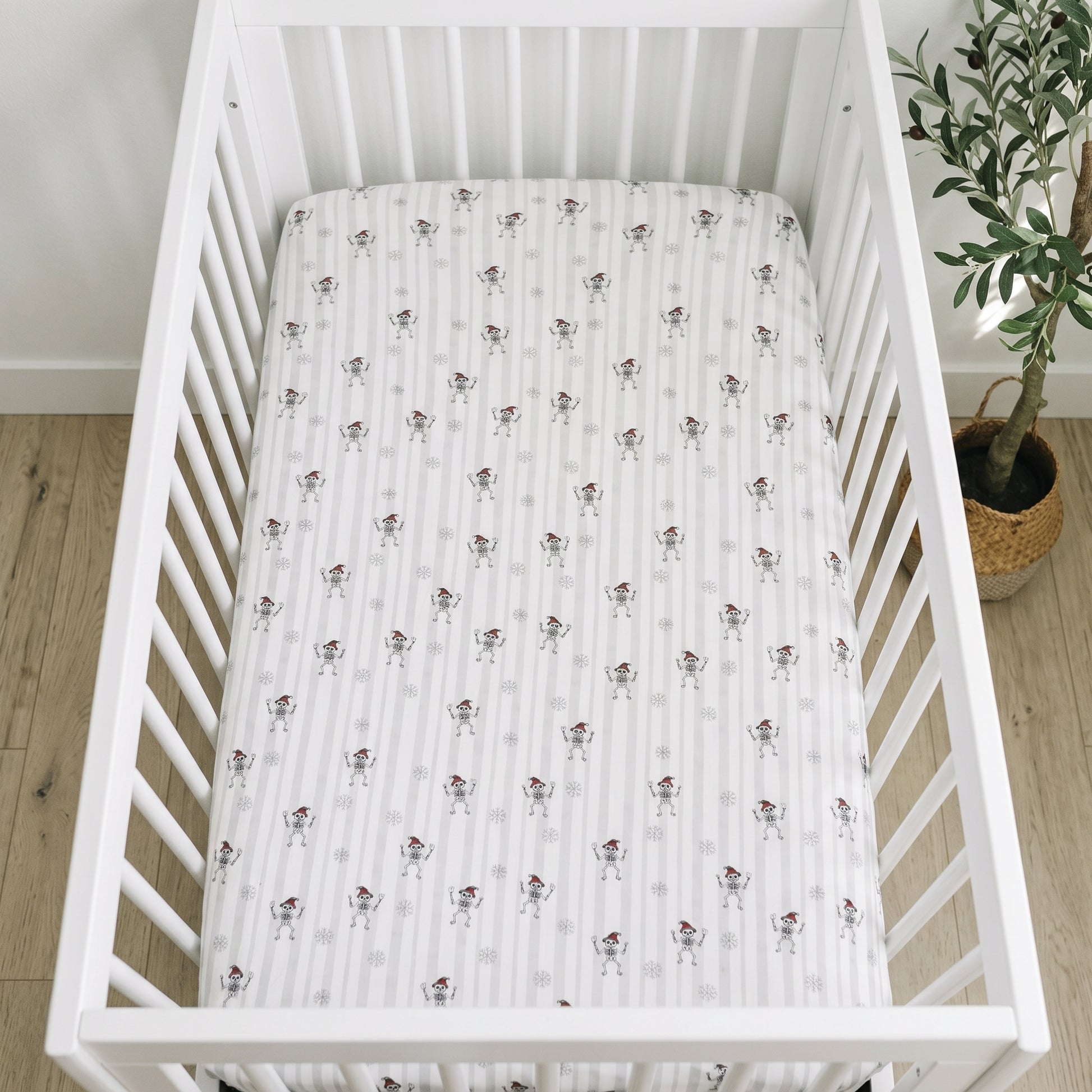 A white baby crib with a fitted sheet featuring a repeating pattern of small cartoon dogs, shown in a nursery setting with light wood flooring and a potted plant.