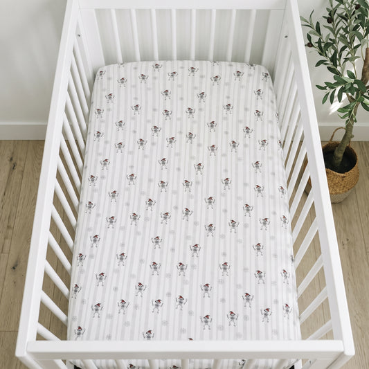 A white baby crib with a fitted sheet featuring a repeating pattern of small cartoon dogs, shown in a nursery setting with light wood flooring and a potted plant.
