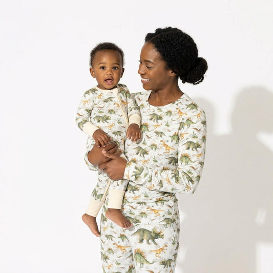 A woman holding a baby, both wearing matching long-sleeve pajamas with an all-over dinosaur print on an off-white background. The pajamas feature green, brown, and orange dinosaurs, with solid cream cuffs and collars.
