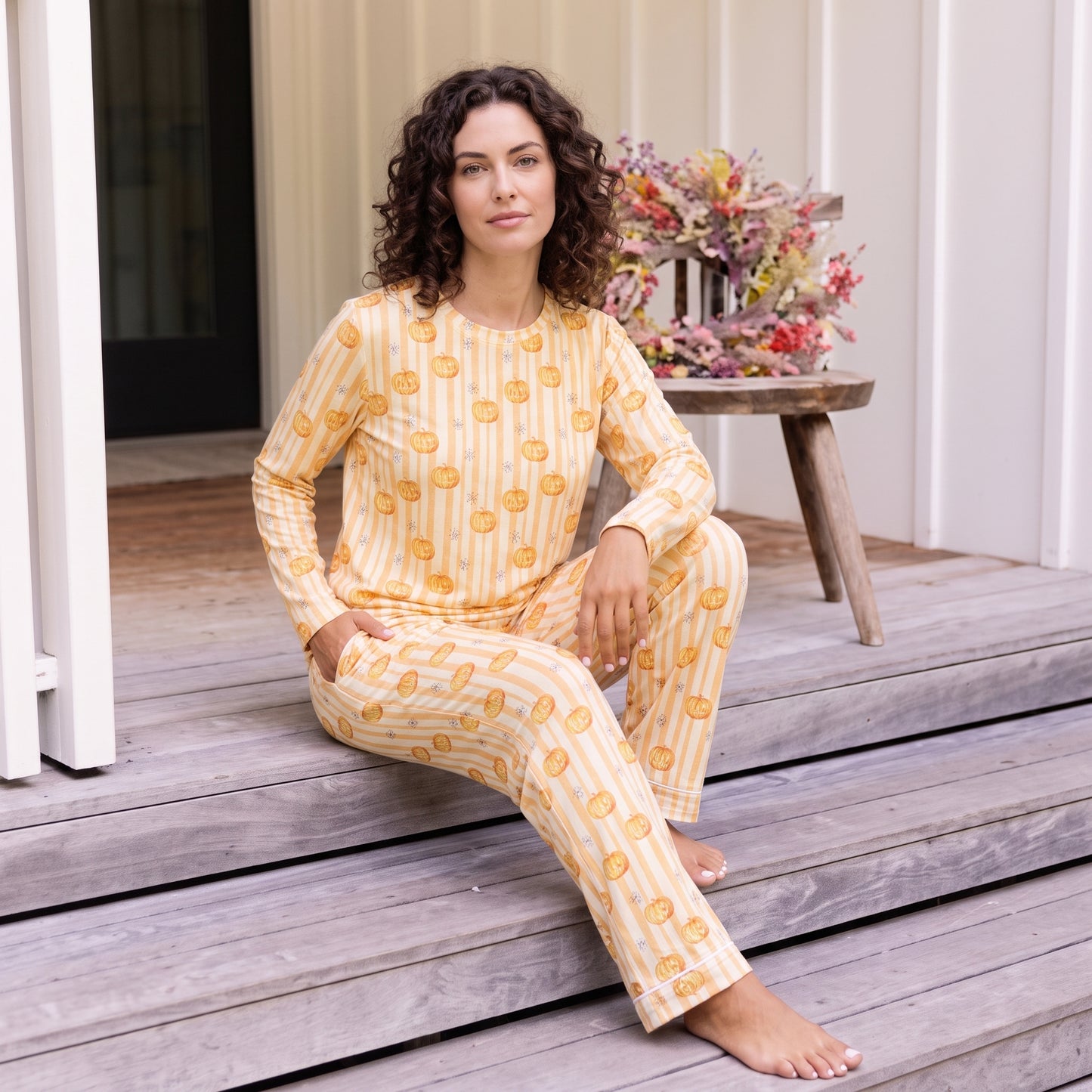 A woman wearing a yellow and white striped pajama set with a yellow sun pattern sits on wooden steps in front of a house, with a small bench and flowers in the background.