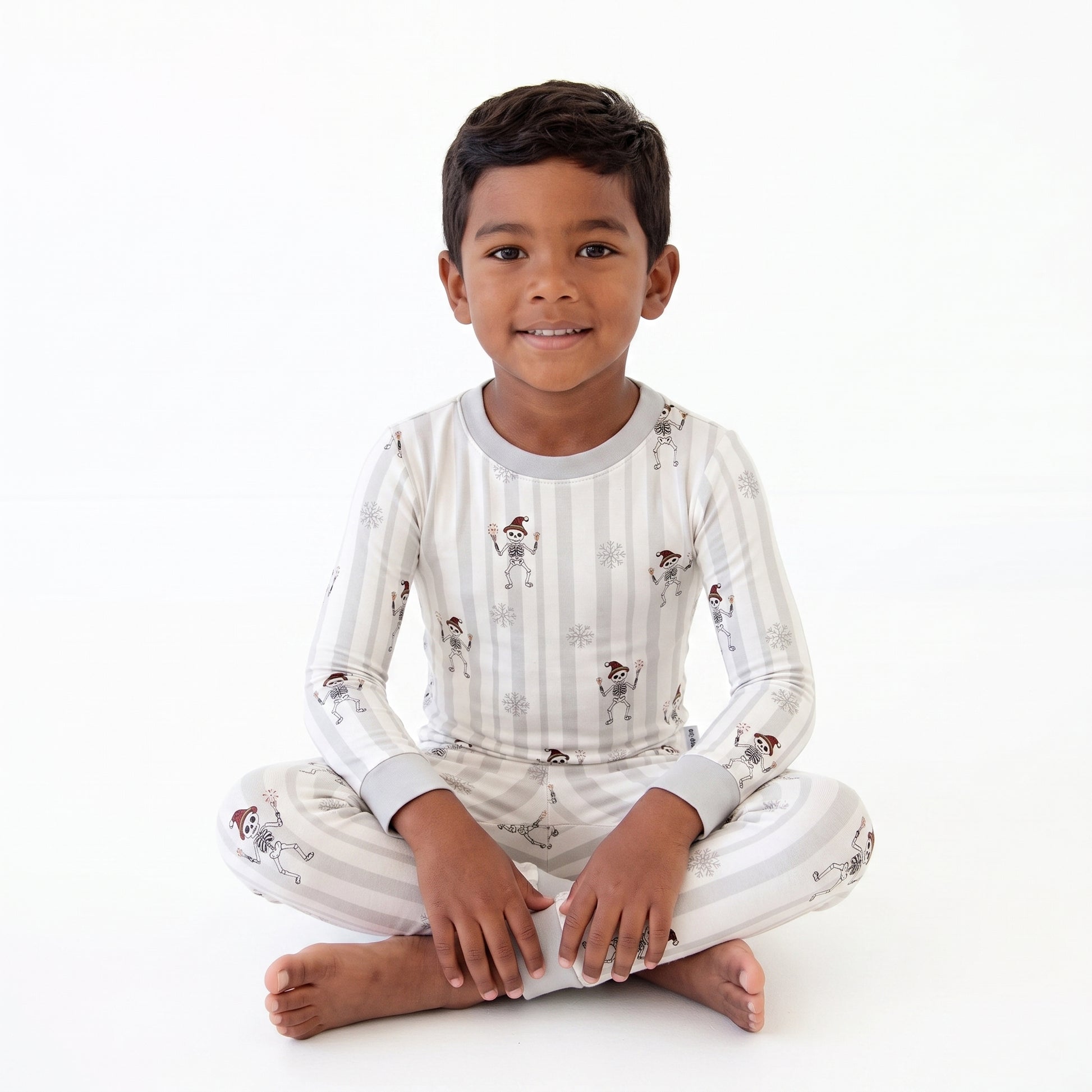 A young boy sitting cross-legged on a white studio background, wearing a long-sleeve pajama top and matching pants with a light gray and white stripe pattern and small astronaut illustrations.