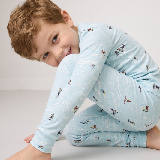 A young boy with curly light brown hair wearing a light blue long-sleeve pajama set with a small penguin print, posing on a light gray studio floor.