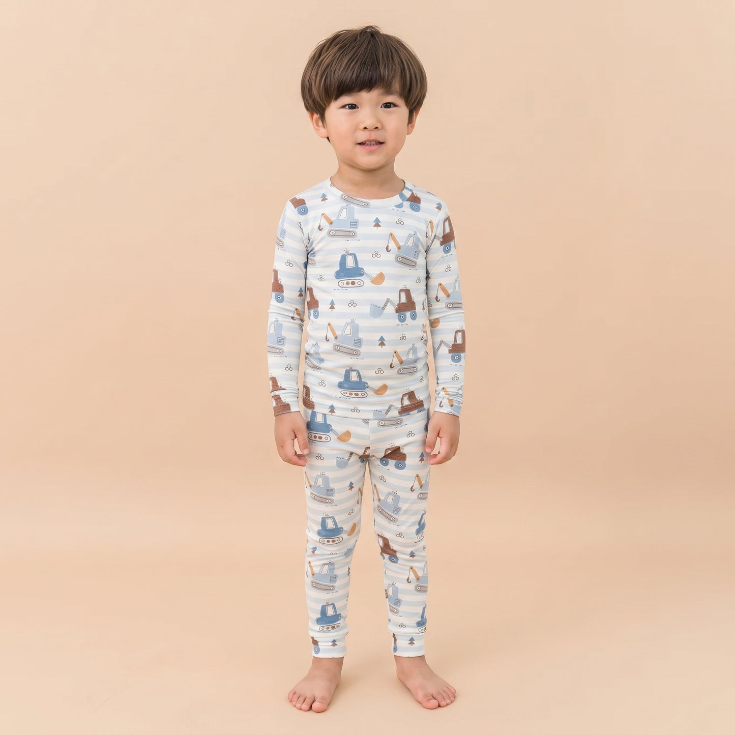 A young child standing barefoot against a solid beige background, wearing a long-sleeve pajama set with a blue and brown construction vehicle pattern on a white base.