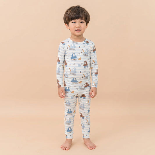 A young child standing barefoot against a solid beige background, wearing a long-sleeve pajama set with a blue and brown construction vehicle pattern on a white base.