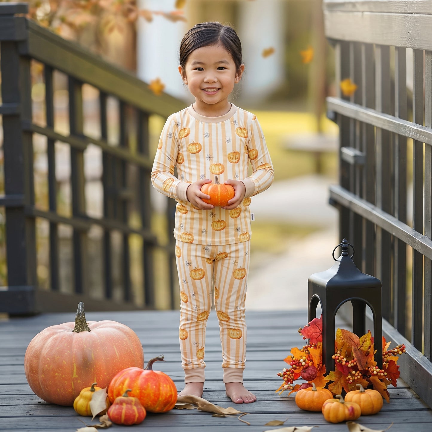 A young child stands barefoot on a wooden deck wearing a cream and orange striped pajama set with pumpkin prints. The child is holding a small orange pumpkin, with pumpkins, autumn leaves, and a black lantern scattered around them.