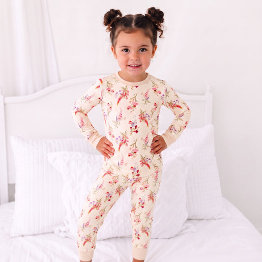 A young girl with her hair in two buns stands on a white bed wearing a cream-colored long-sleeve pajama set with a red and green floral print.