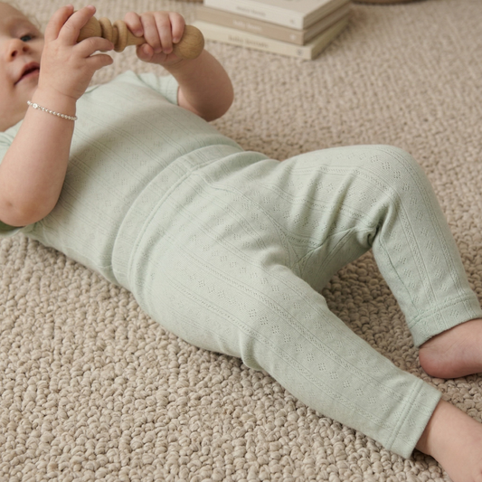 Baby in a light green outfit lying on a beige carpet, holding a wooden toy.