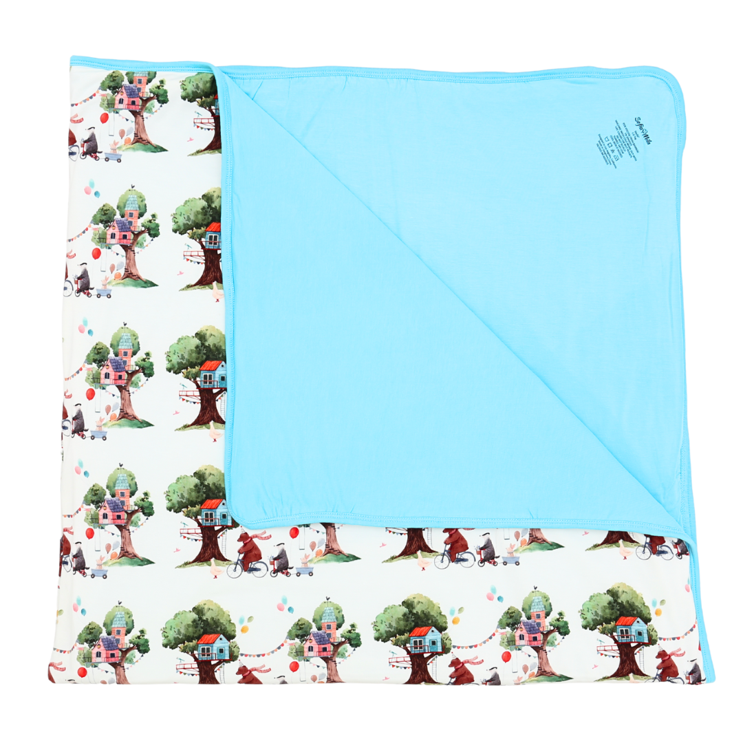Children's blanket with tree house pattern on one side and solid blue color on the other, displayed against a white background.